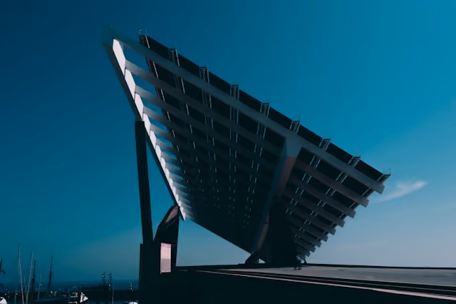 Large solar panel tilted upwards against a clear blue sky near a body of water.
