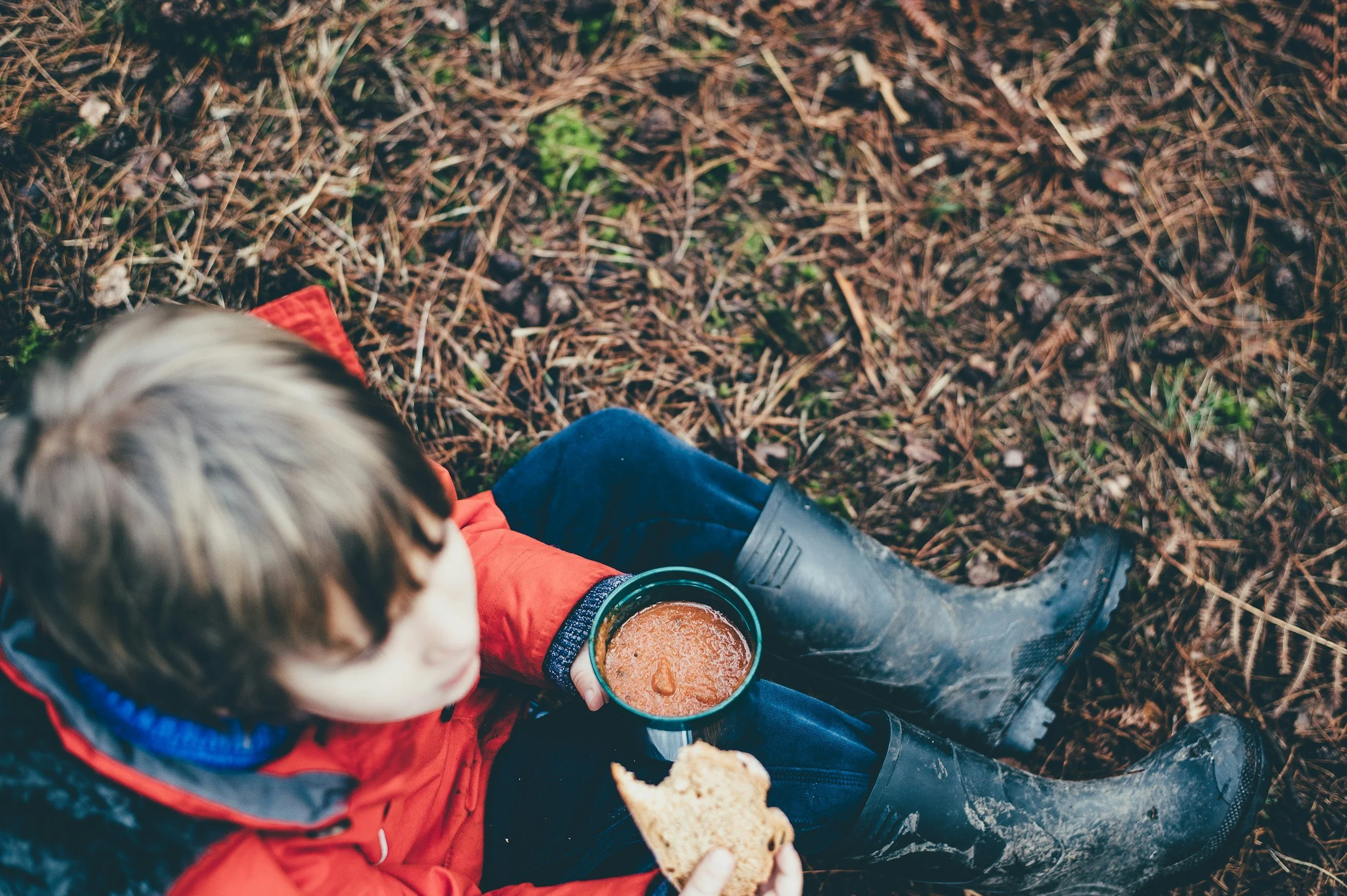A boy in a red jacket sitting outdoors holding a mug of hot cocoa