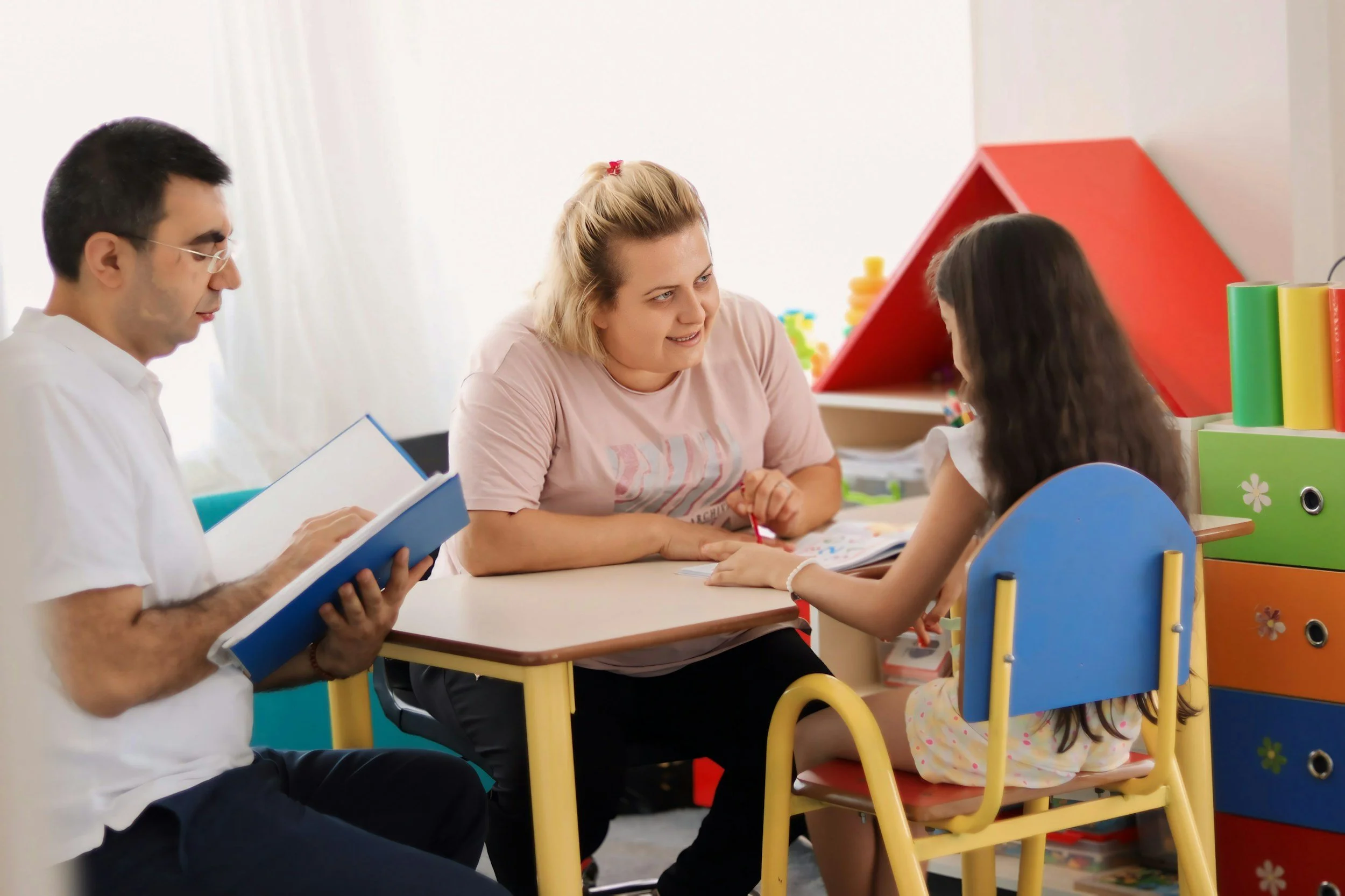 Two adults sitting at a small table with a young girl in a colorful playroom
