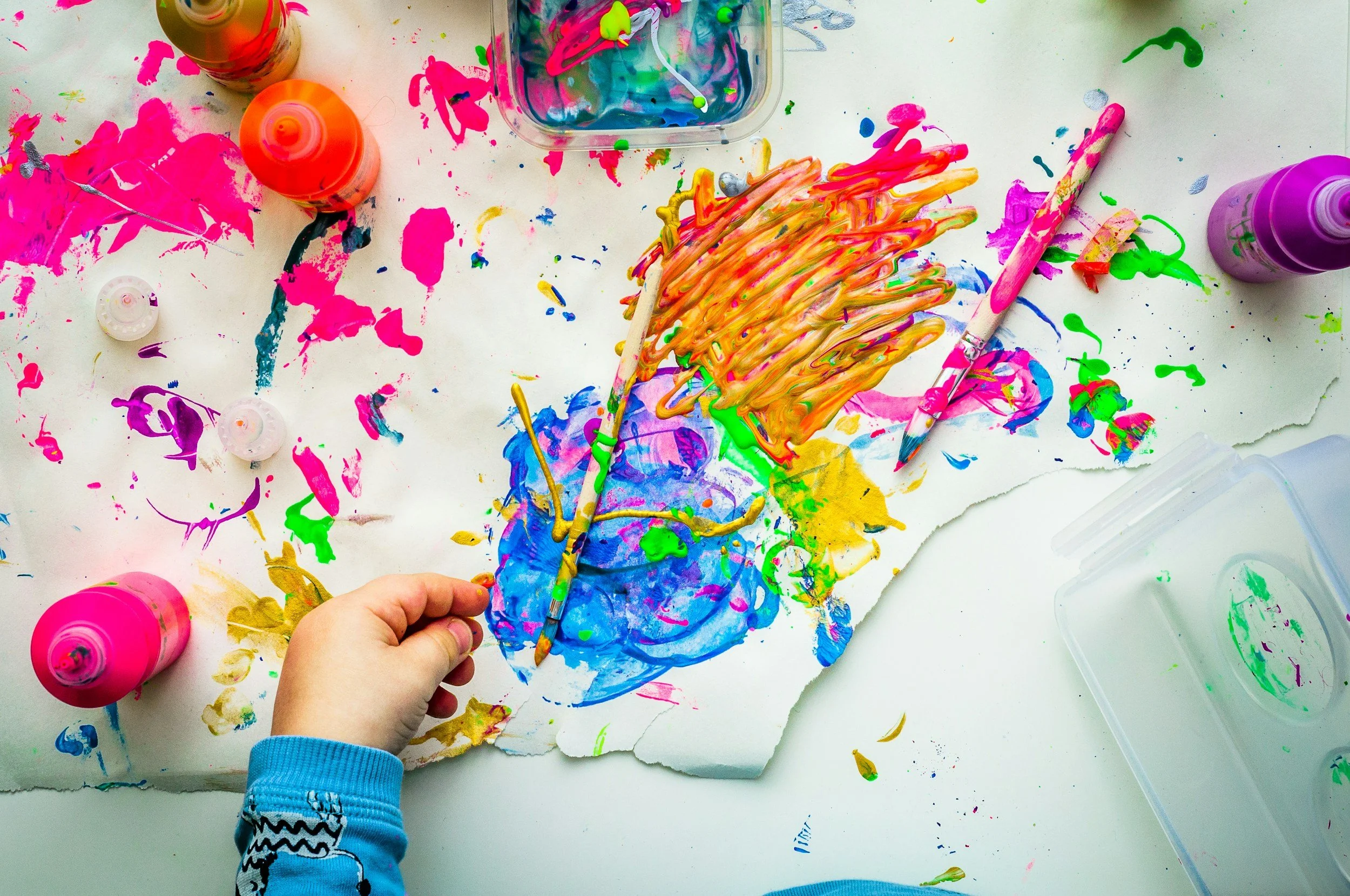 A child's hand painting with bright colors on a paint-splattered table