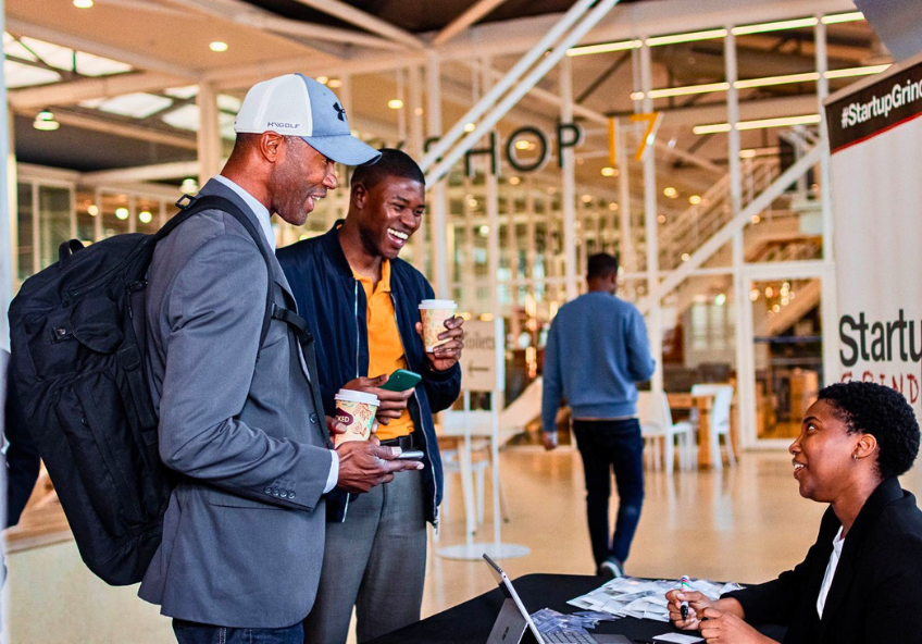Encontro de comunidade no evento da Startup Grind no Workshop 17, no Waterfront em Cape Town. Em primeiro plano, dois membros conversam com copos de café, destacando-se Geovany (fundador da Comoeu) e Giovanni Freeman, também conhecido como Coach Gio (fundador da G3D e da associação AGH African Grassroot Hoops), enquanto a equipa de acolhimento faz o check-in na banca de receção.