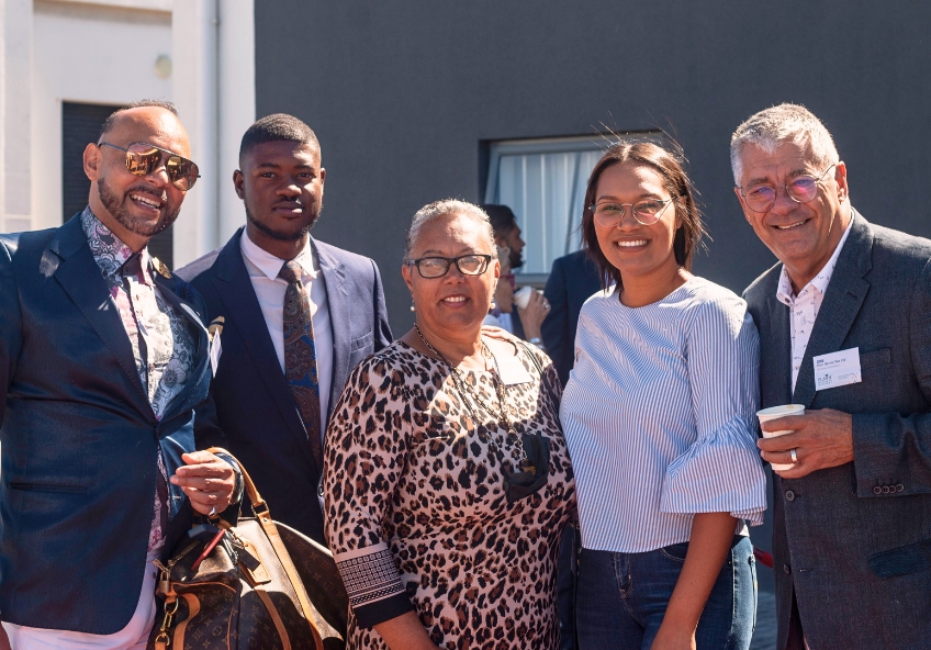 Fotografia de grupo num evento de networking organizado pela Comoeu na África do Sul. Ao centro e à esquerda, o fundador Geovany, de fato azul, posa ao lado de líderes, investigadores e membros da equipa da Ed Bham Foundation. O grupo de cinco profissionais sorri para a câmara num encontro focado em usar a tecnologia para democratizar o acesso à educação nas comunidades.