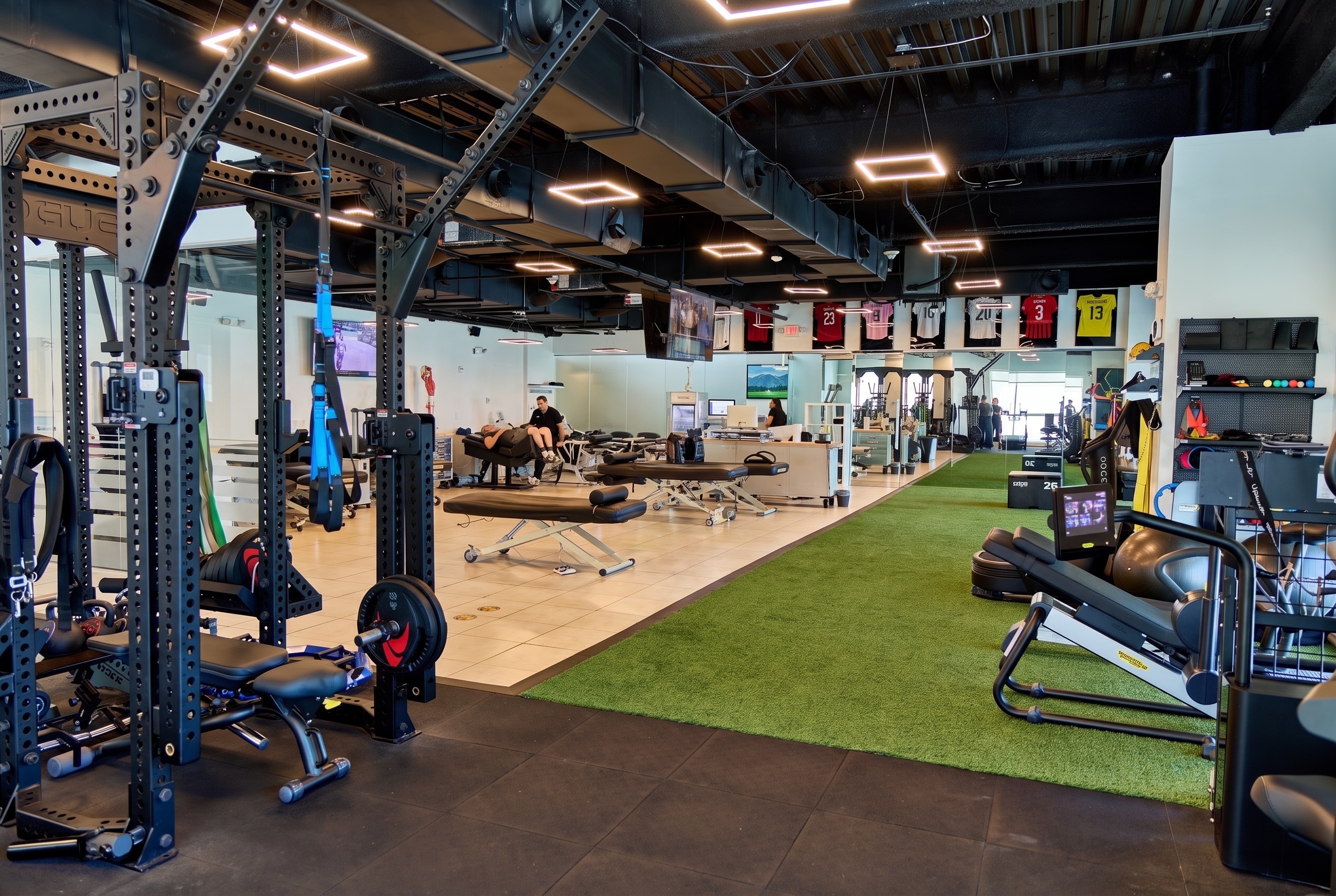 Modern gym with weight racks, exercise benches, green turf, treadmills, and sports jerseys on the wall.