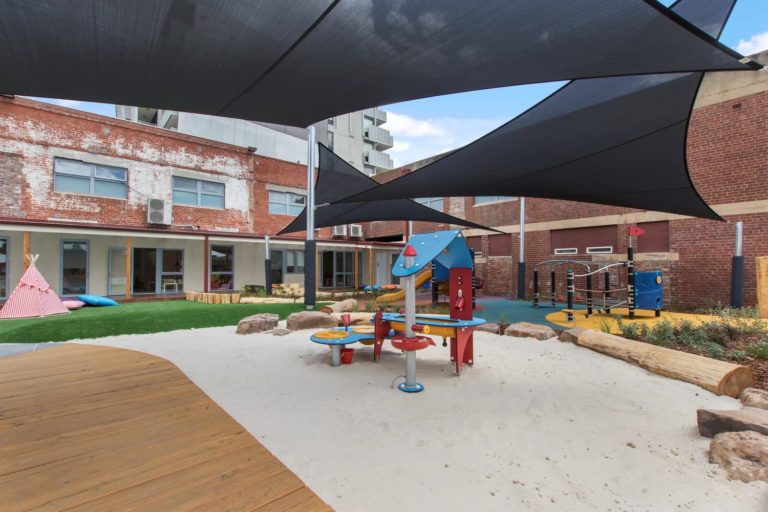 Modern outdoor playground with sand, shade sails, climbing equipment, and seating near brick buildings.
