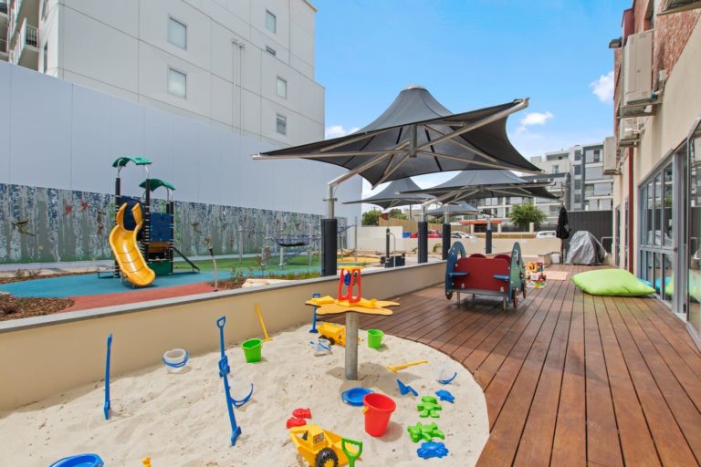 Outdoor playground with sandpit, colorful toys, slide, and shaded seating on wooden deck.