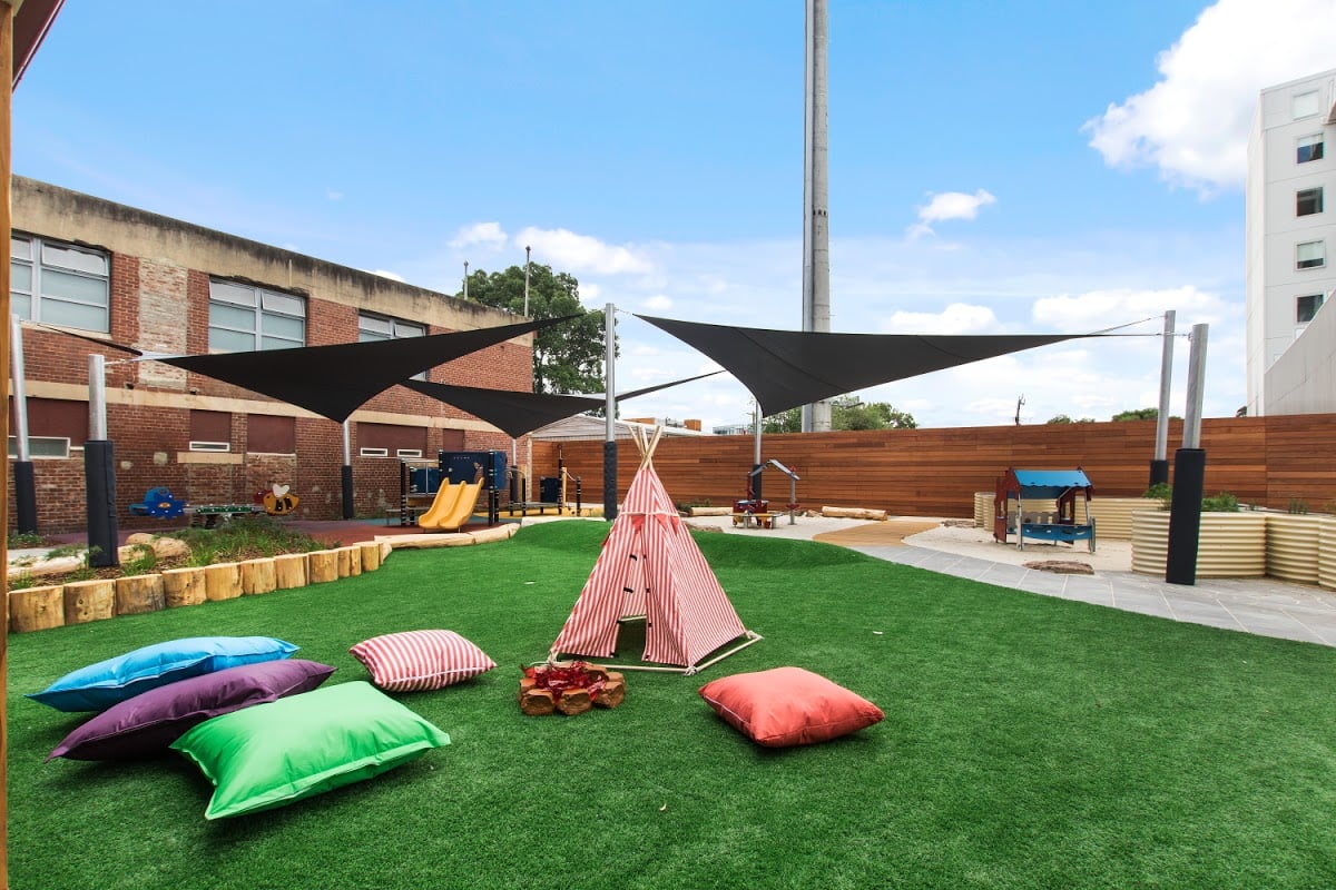 Outdoor play area with green grass, colorful pillows, a striped teepee, and playground equipment under shade sails.