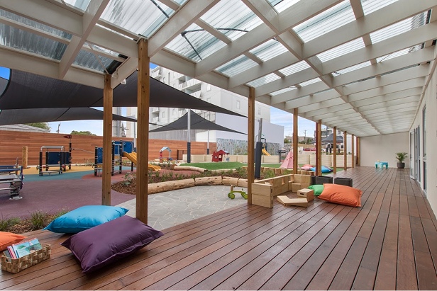 Covered wooden deck with cushions and wooden toys overlooking a colorful playground with shade sails.