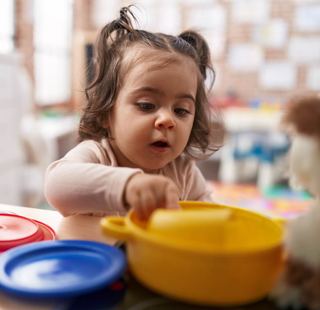Toddler with pigtails playing with a yellow toy pot and colorful lids at a table indoors.