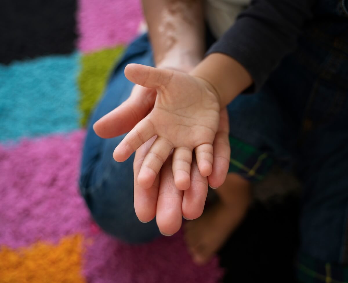Small child's hand resting open on an adult's open hand over a colorful patchwork rug.