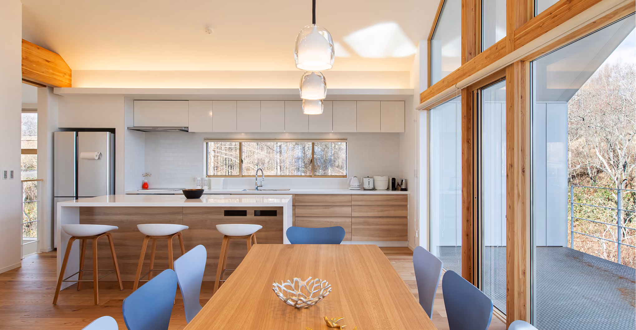 Modern kitchen with wood and white cabinets, island with stools, and large windows overlooking a balcony.