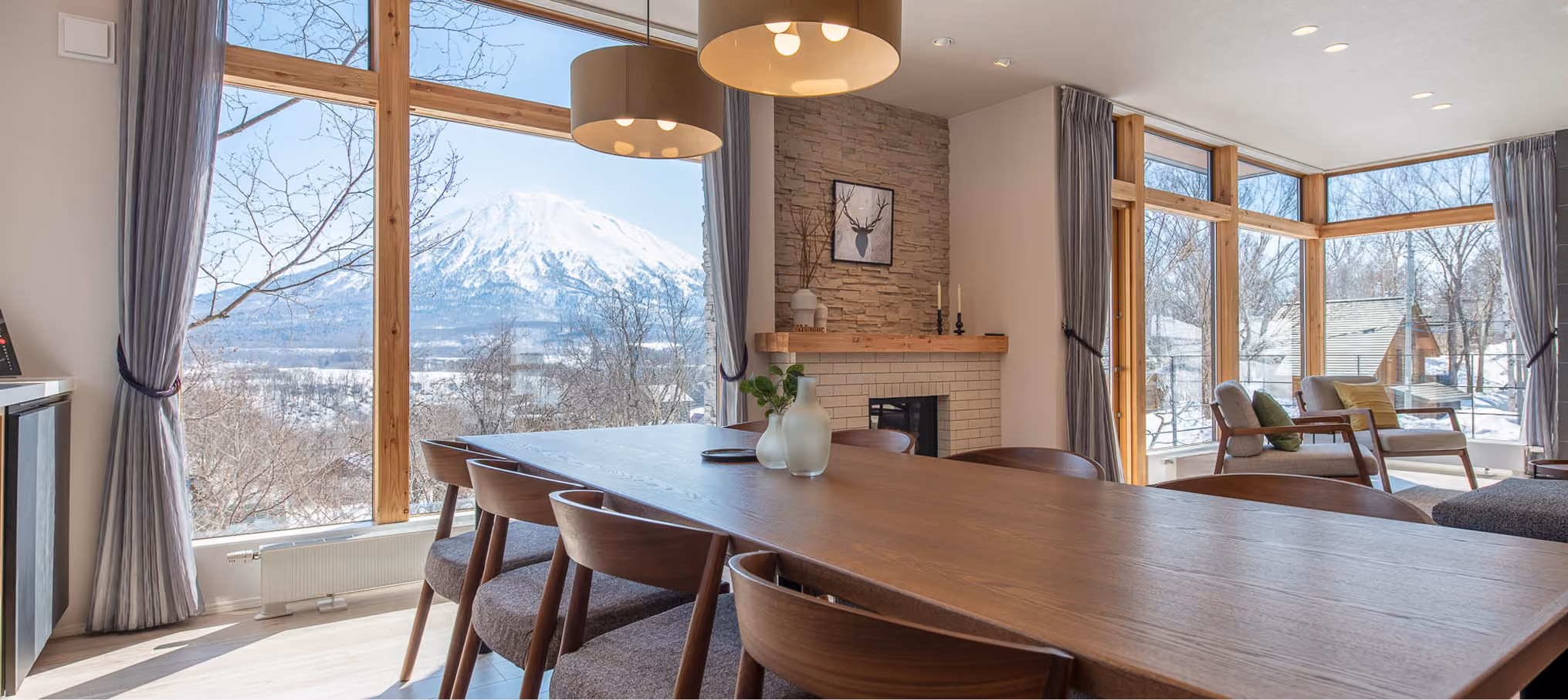 Dining room with wooden table, chairs, large windows showing snowy mountain and trees outside.
