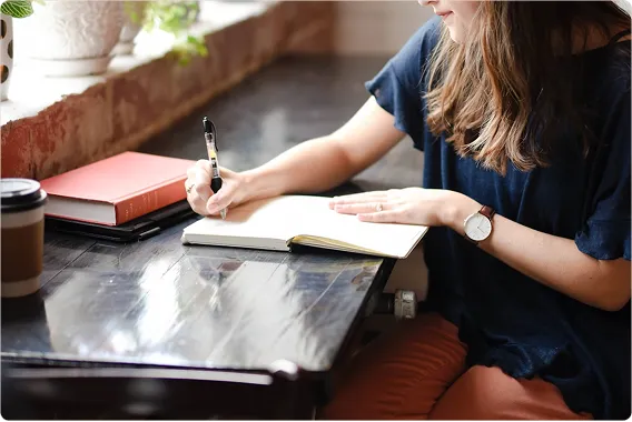 Person writing and reflecting in a notebook near a window