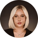 Blonde Frau mit Brille und rotem Lippenstift, trägt Kette, vor dunklem Hintergrund.