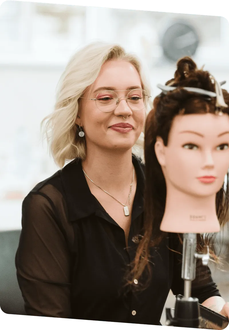 Friseurin mit blonden Haaren und Brille stylt Haare an Friseurkopf im Salon.
