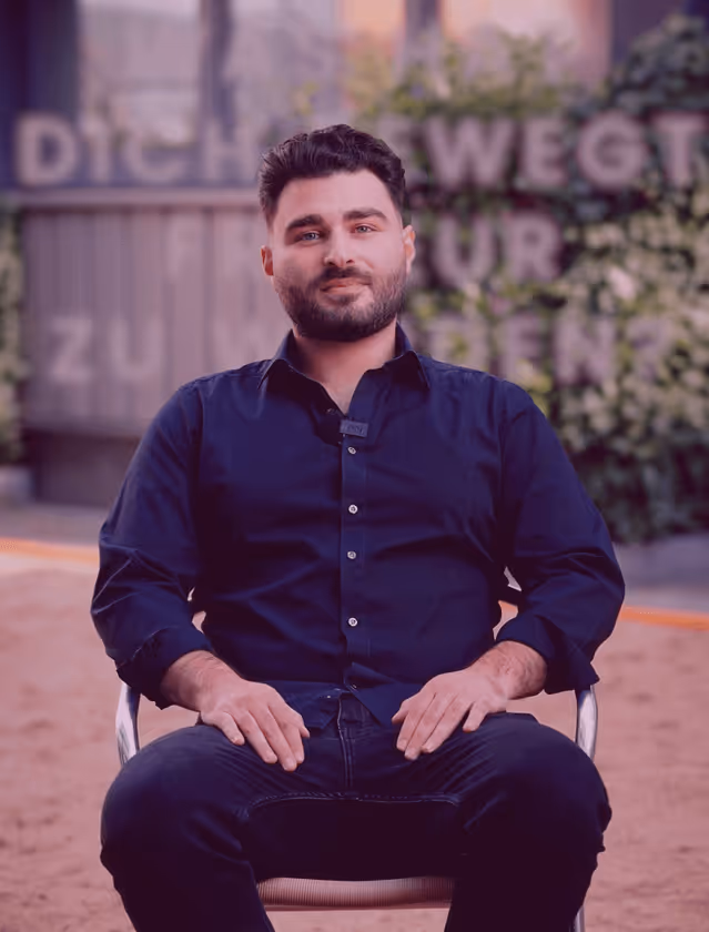 Person mit dunklem Hemd sitzt auf Stuhl vor unscharfem Hintergrund mit Pflanzen und Schrift.