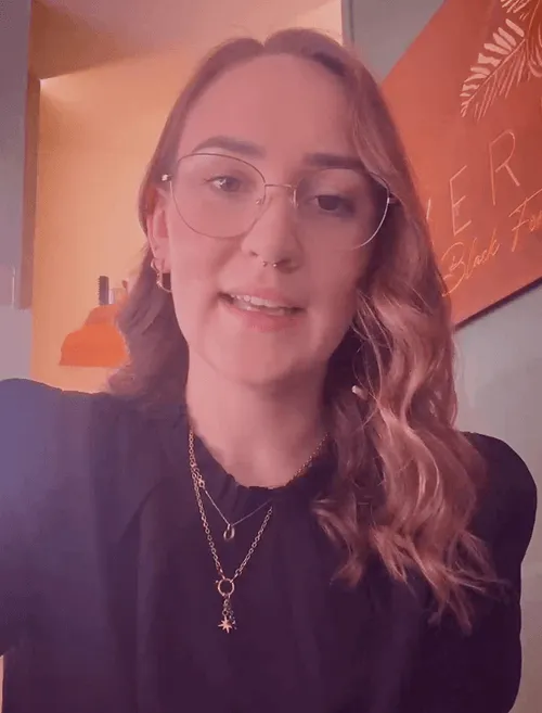 Frau mit Brille und welligem Haar spricht in die Kamera, trägt schwarze Bluse, Kette; orangefarbene Wand im Hintergrund.