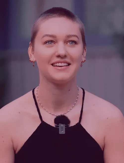 Lächelnde Person mit kurzem Haar, trägt ein schwarzes Oberteil und ein Mikrofon, im Freien stehend.