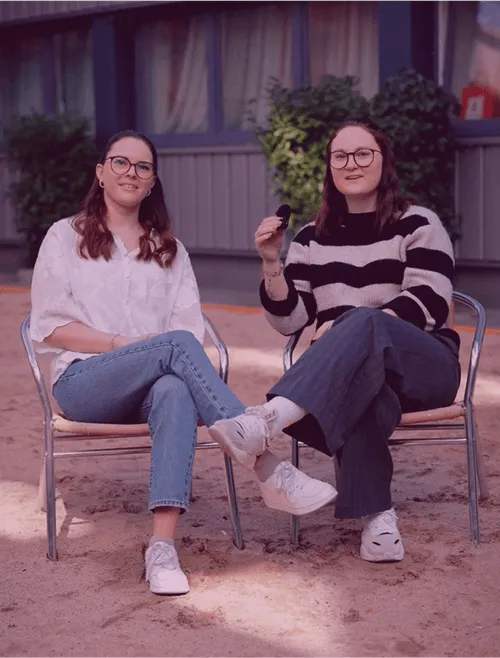 Zwei lächelnde Frauen sitzen draußen auf Stühlen, tragen Jeans und Brillen, umgeben von Pflanzen und einem Gebäude.