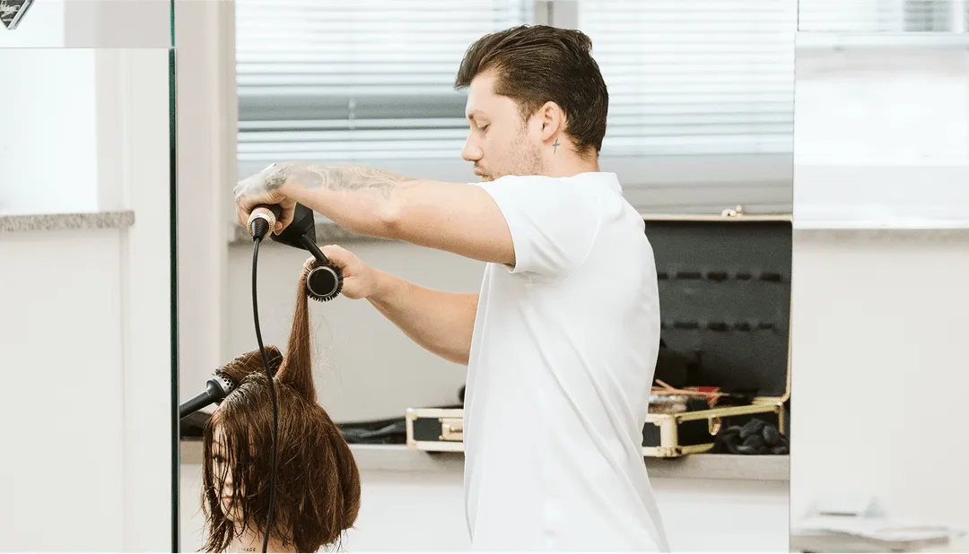 Friseur stylt kundinnes nasses Haar mit Föhn und Rundbürste in modernem Salon.