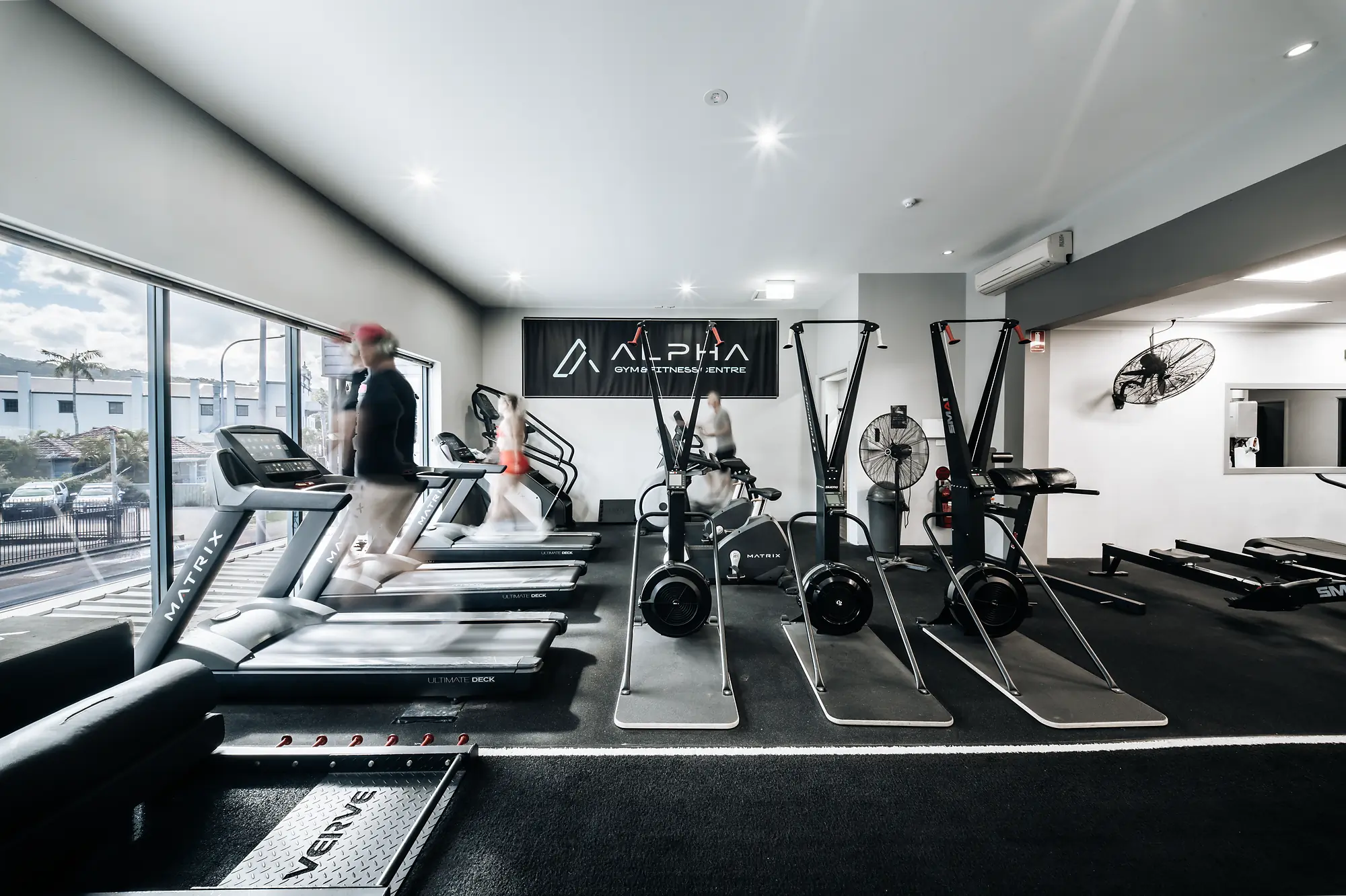 Gym interior with treadmills, exercise bikes, and fitness machines under bright ceiling lights.