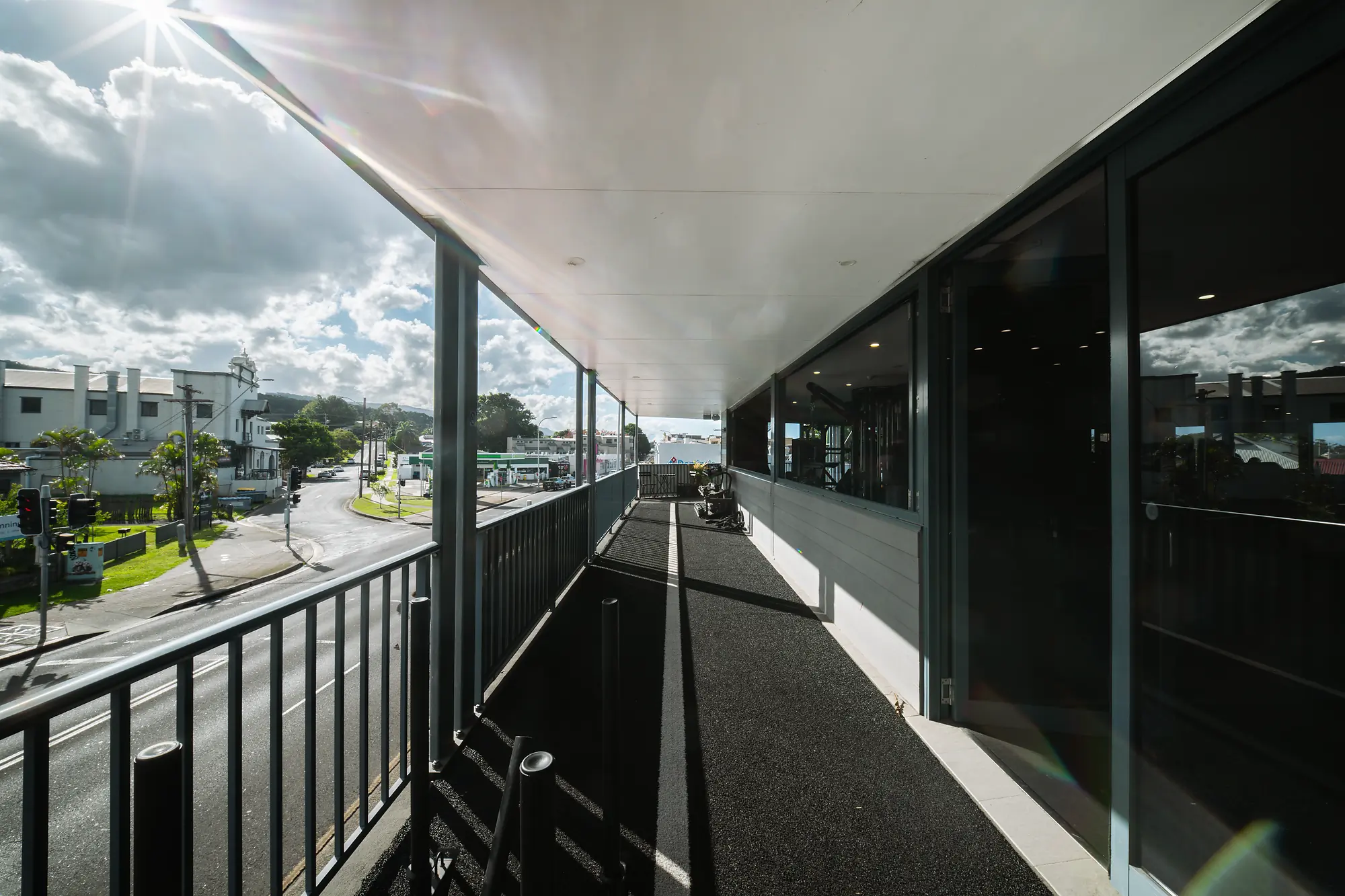 Sunlit outdoor balcony with black railing overlooking a street and buildings under a cloudy sky.