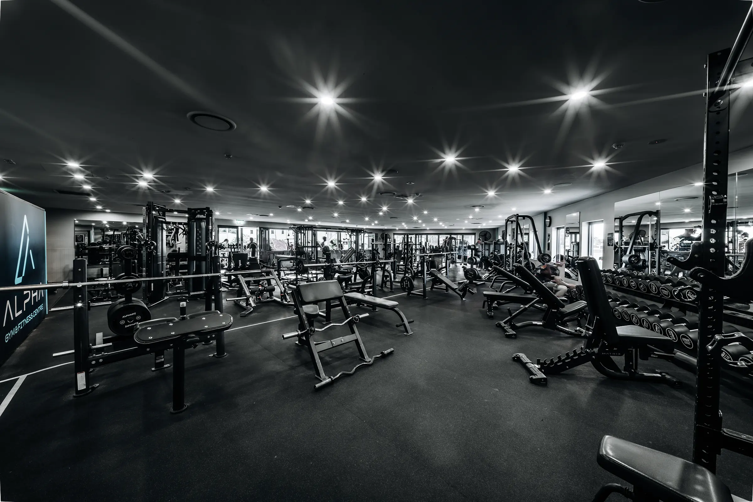 Spacious gym with weight benches, dumbbells, machines, and bright ceiling lights.