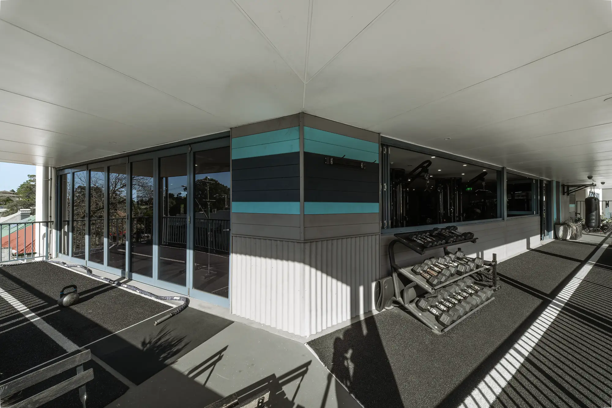 Outdoor gym corner with kettlebell, battle rope, dumbbells, and punching bag under covered roof.