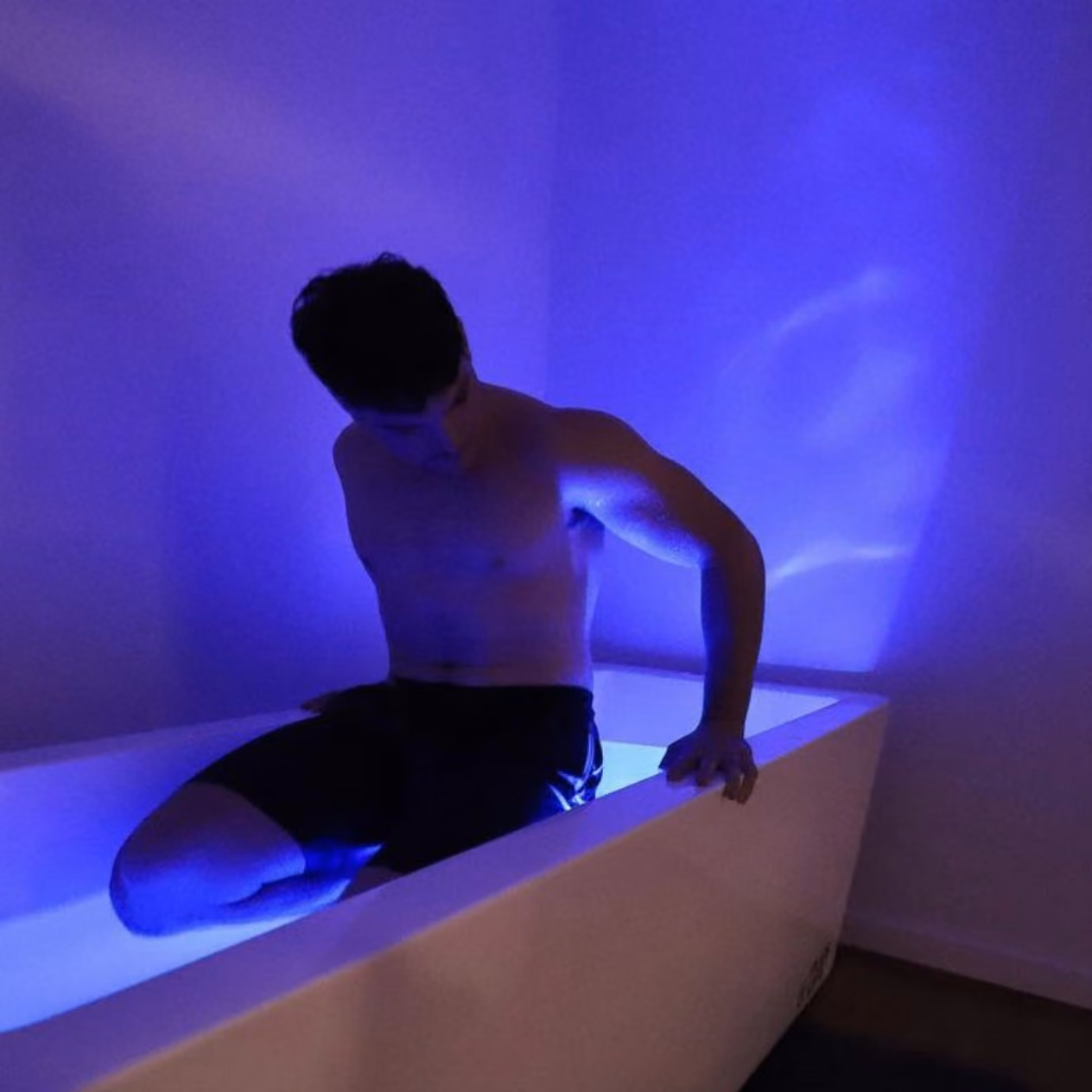 Shirtless man in black shorts sitting on the edge of a tub illuminated by blue light.