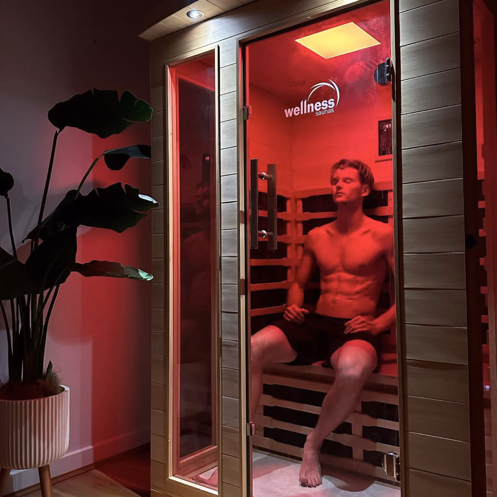 Shirtless man sitting and relaxing inside a wooden infrared sauna with red lighting.