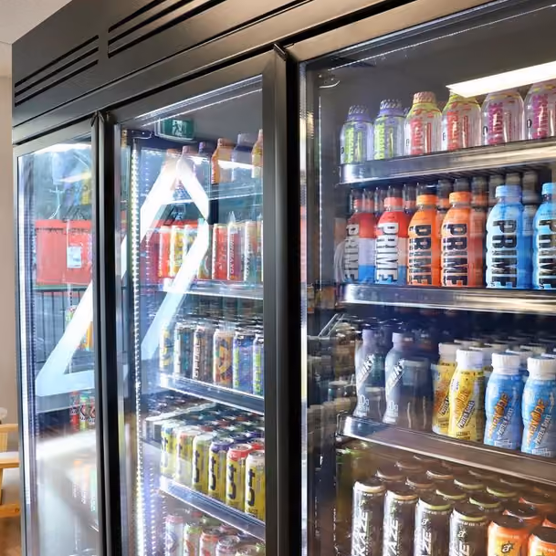 Refrigerator with glass doors stocked with various colorful supplement drink bottles and cans on shelves.