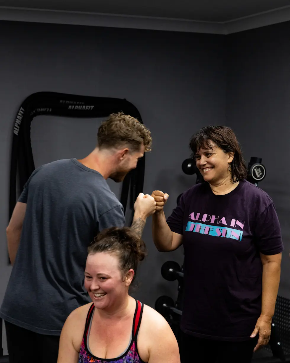 Two people fist bump during a gym session while a smiling woman sits in front of them.
