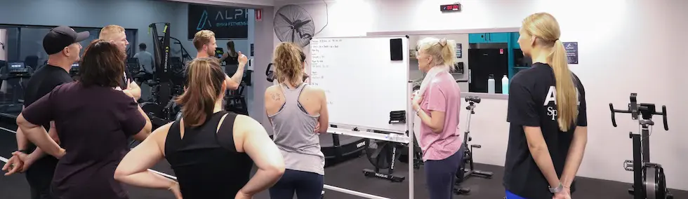 Group of people in gym standing around a whiteboard with workout instructions.