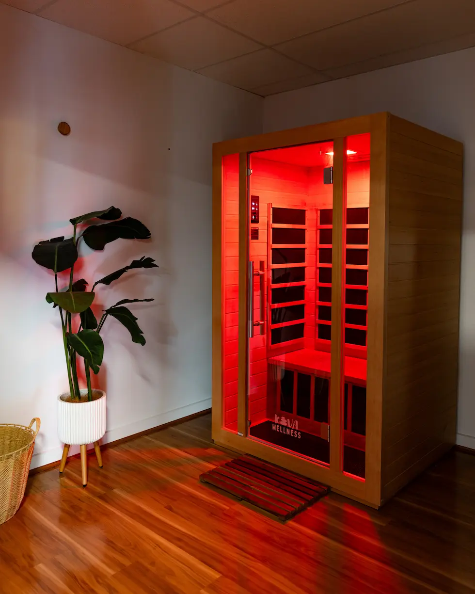 Wooden infrared sauna cabin with red interior light beside potted plant in a room with hardwood floor.