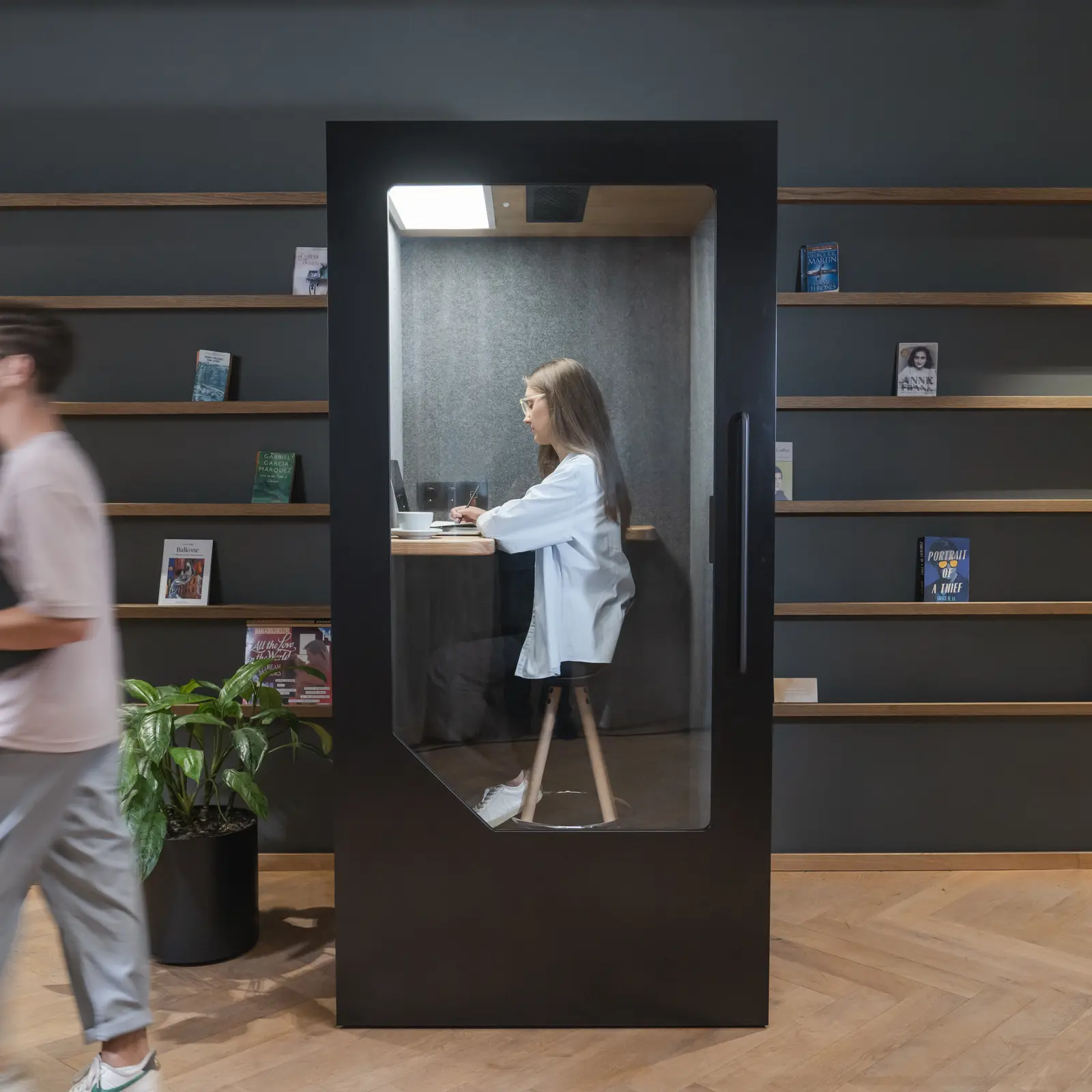 Person_working_inside_a_dark_acoustic_office_pod_with_colleague_walking_past_in_open_office