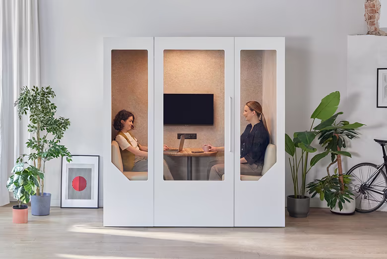 Two women seated facing each other inside a modern white office pod with large windows, surrounded by indoor plants and a bicycle in a bright room.