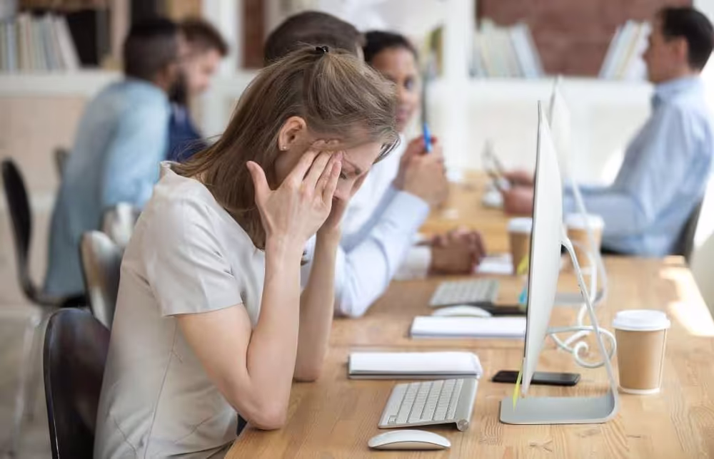 Woman sitting at office desk holding her head in frustration with blurred colleagues working in the background.
