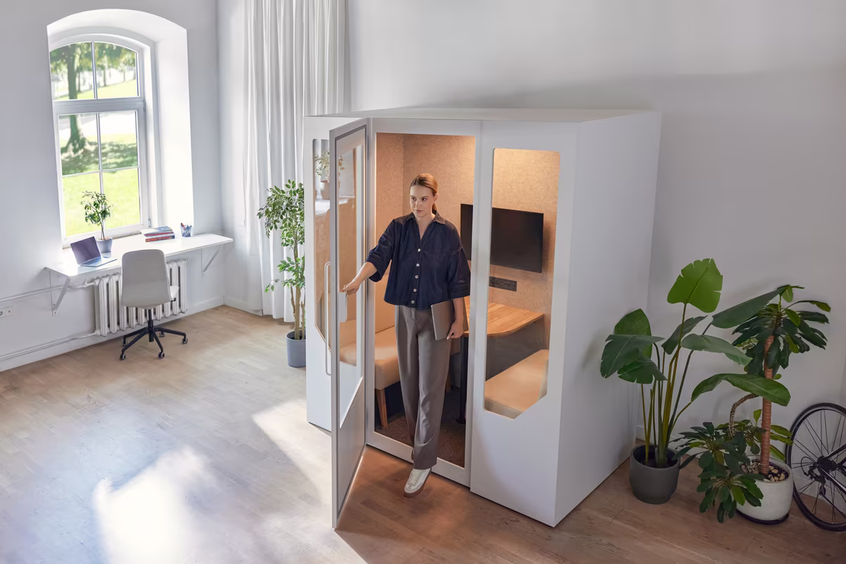 Person taking a phone call inside a soundproof office booth within an open workspace