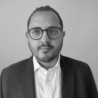 Man with dark hair and beard wearing glasses, white shirt, and dark blazer against plain background.