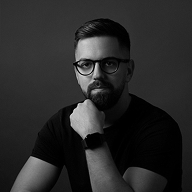 Black and white portrait of a man with glasses, beard, and smartwatch resting chin on hand.