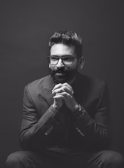 Man with glasses and beard in a suit sitting with hands clasped, looking contemplative against a dark background.