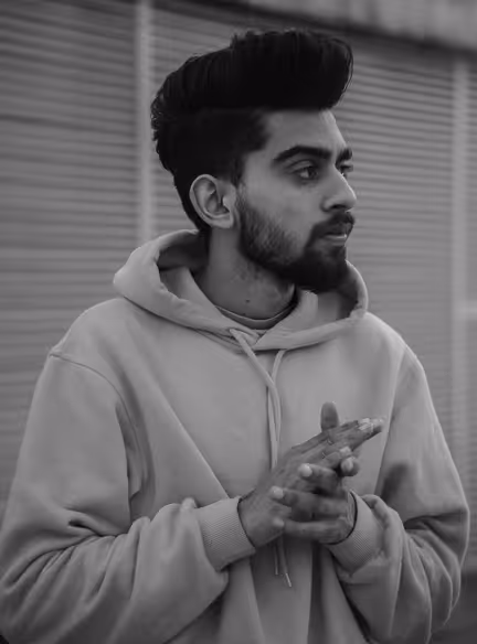 Man with styled hair and beard wearing a hoodie, looking to the right, hands clasped near chest.