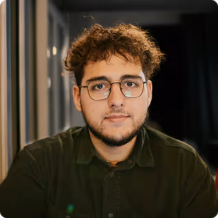 Young man with curly hair and glasses wearing a dark green shirt, looking at the camera indoors.
