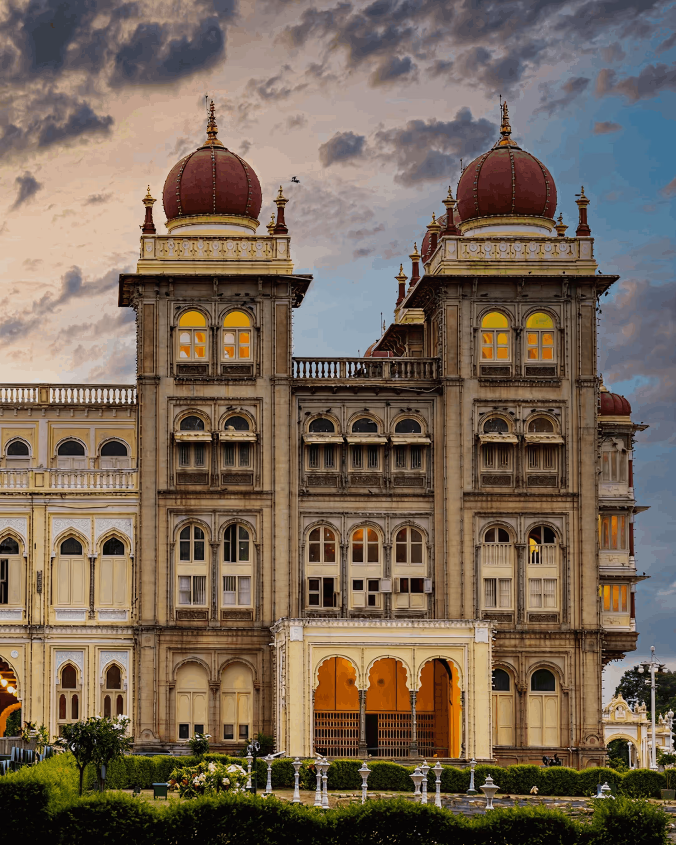 Historic palace with two large red domes, arched windows, and illuminated interiors under a cloudy sky.