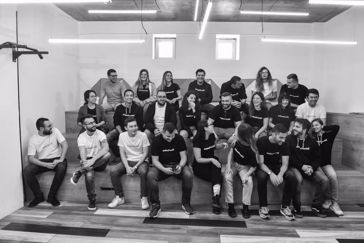 Group of 23 people sitting on wooden steps in a modern room, wearing matching black and white shirts.