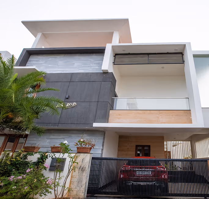 Modern two-story house with a balcony, black gate, plants, and a red car parked inside.