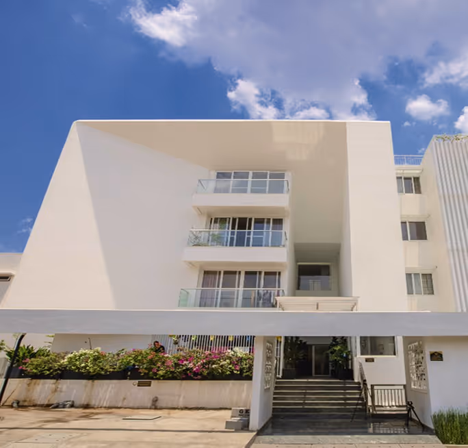 Modern white multi-story building with balconies, stairs at entrance, under a partly cloudy blue sky.