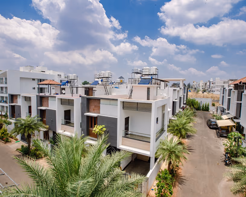 Modern white apartment buildings with balconies, palm trees, cars, and a partly cloudy sky.