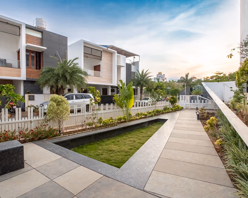 Modern residential area with paved walkway, manicured lawn, palm trees, and contemporary houses under blue sky.