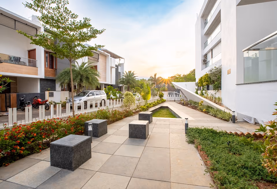 Modern residential street with greenery, stone benches, palm trees, and setting sun in the background.