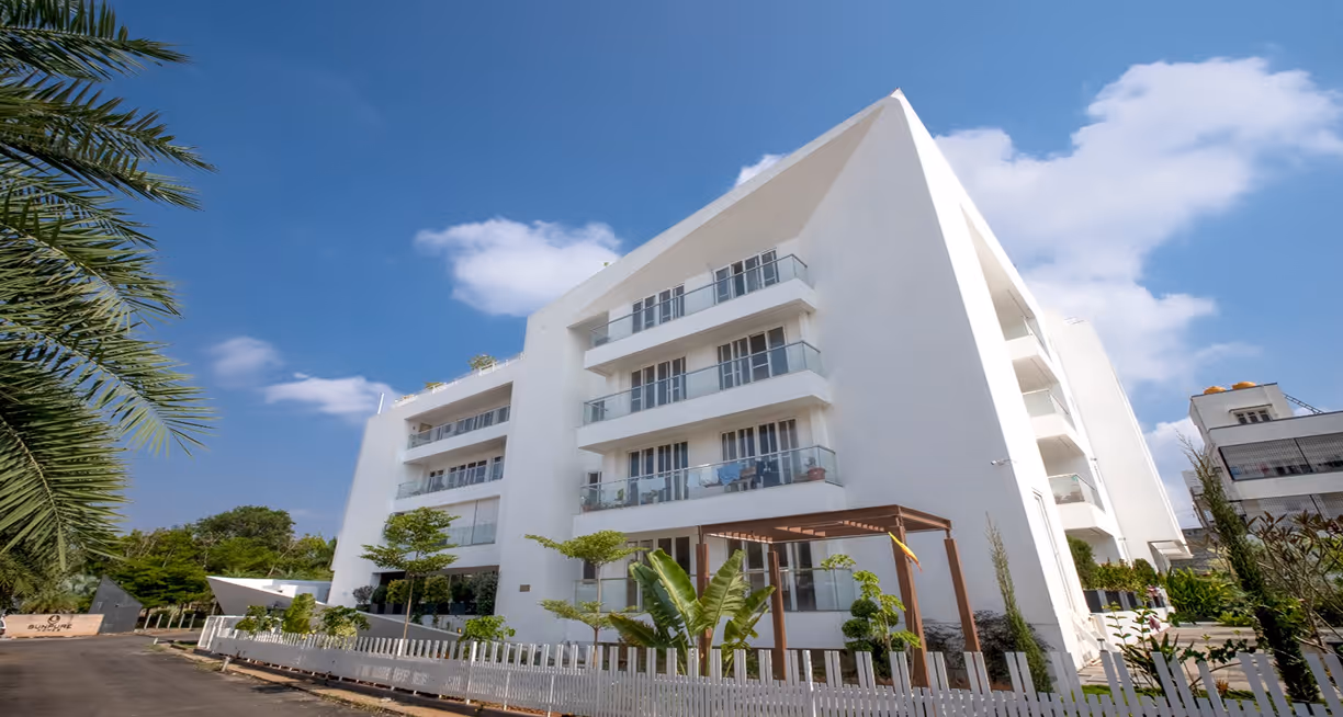 Modern white apartment building with balconies under a blue sky and surrounding green plants.